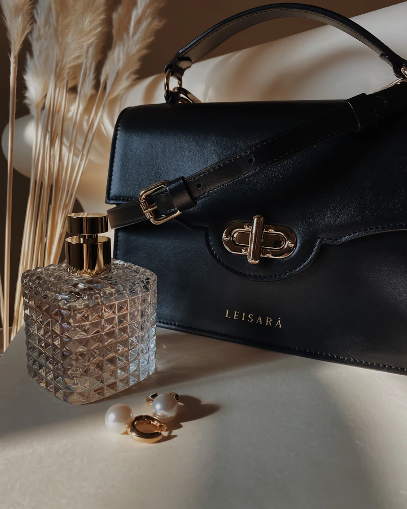 Black leather handbag with gold clasp beside a perfume bottle