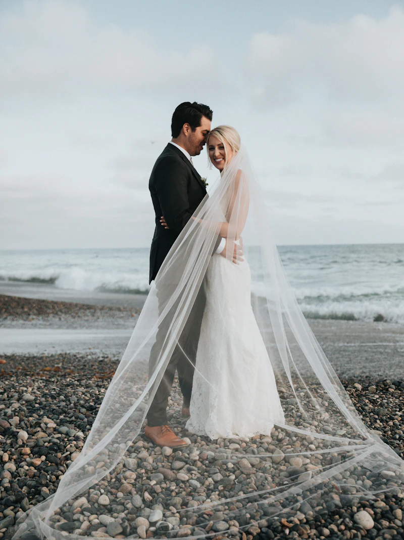 Sarah in a long veil embracing James on a pebble beach by the sea